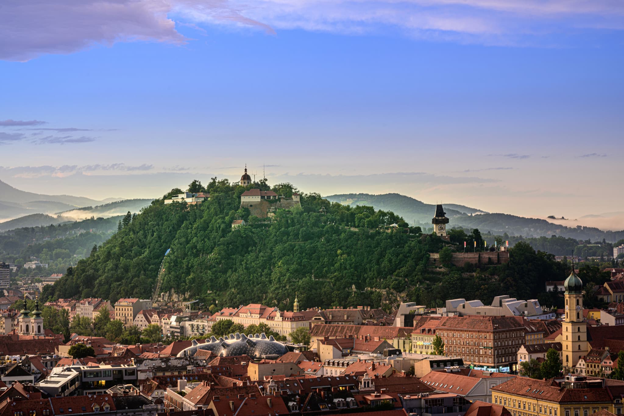 Mittelalterliche Burg auf bewaldeter Anhöhe Mittelalterliche Burg auf einer Anhöhe, umgeben von dichtem Wald, mit Blick auf eine historische Stadt mit roten Ziegeldächern und Kirchturmspitzen in einem Bergtal.