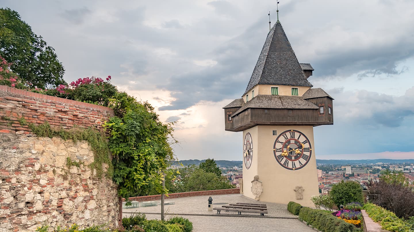 Historic Clock Tower Overlooking City Historic clock tower with a pointed roof and wooden balcony overlooking a city landscape, with ivy-covered stone walls and flowering plants nearby.