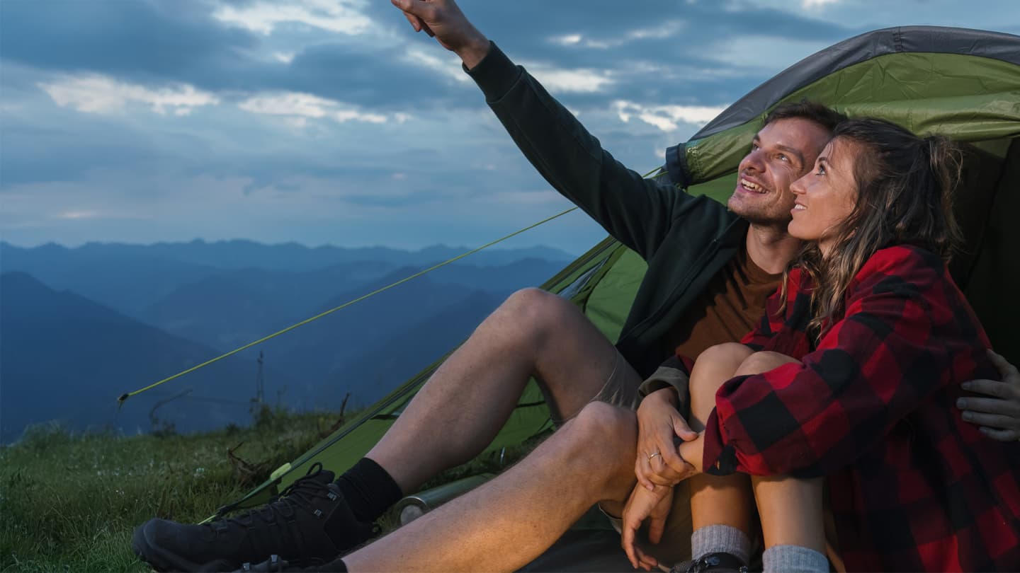 Romantic Mountain Camping Adventure at Sunset Happy couple sitting by green tent on mountain summit pointing at sky during sunset