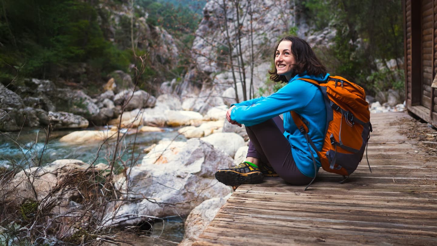 Hiker Resting by Mountain River Smiling female hiker with orange backpack resting by a mountain river