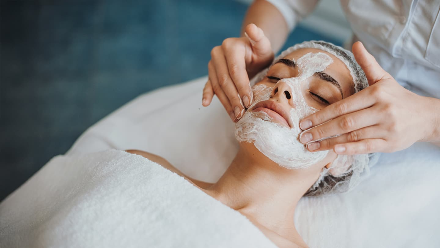 Professional Spa Facial Treatment Esthetician applying cream face mask to relaxed woman at spa