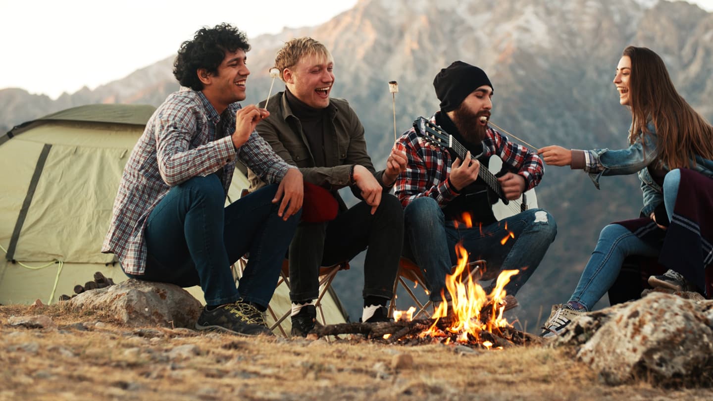 Friends Camping Around Mountain Campfire Four friends laughing around campfire roasting marshmallows with guitar in mountains