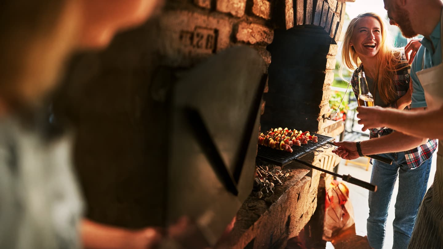 Friends Enjoying Summer Barbecue Party Smiling blonde woman enjoying summer barbecue with friends grilling colorful vegetable kebabs on brick grill
