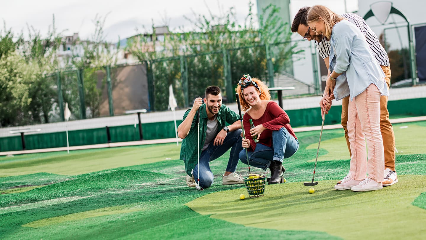 Group Playing Mini Golf Indoors Four people playing mini golf on an indoor course with green barriers and a ball collection basket.