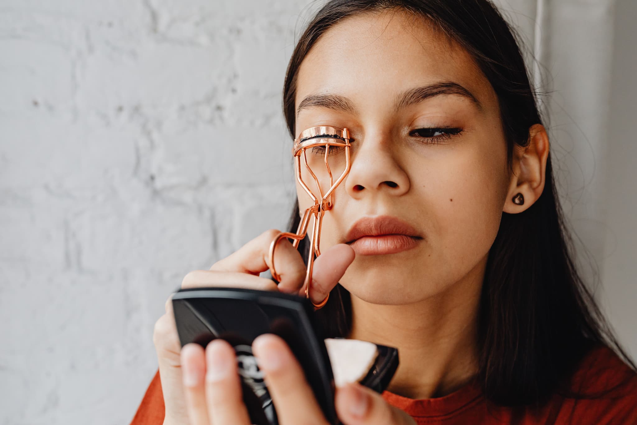 Eyelash Curler and Cosmetic Tool Person holding an orange eyelash curler and a black cosmetic device against a white textured wall.