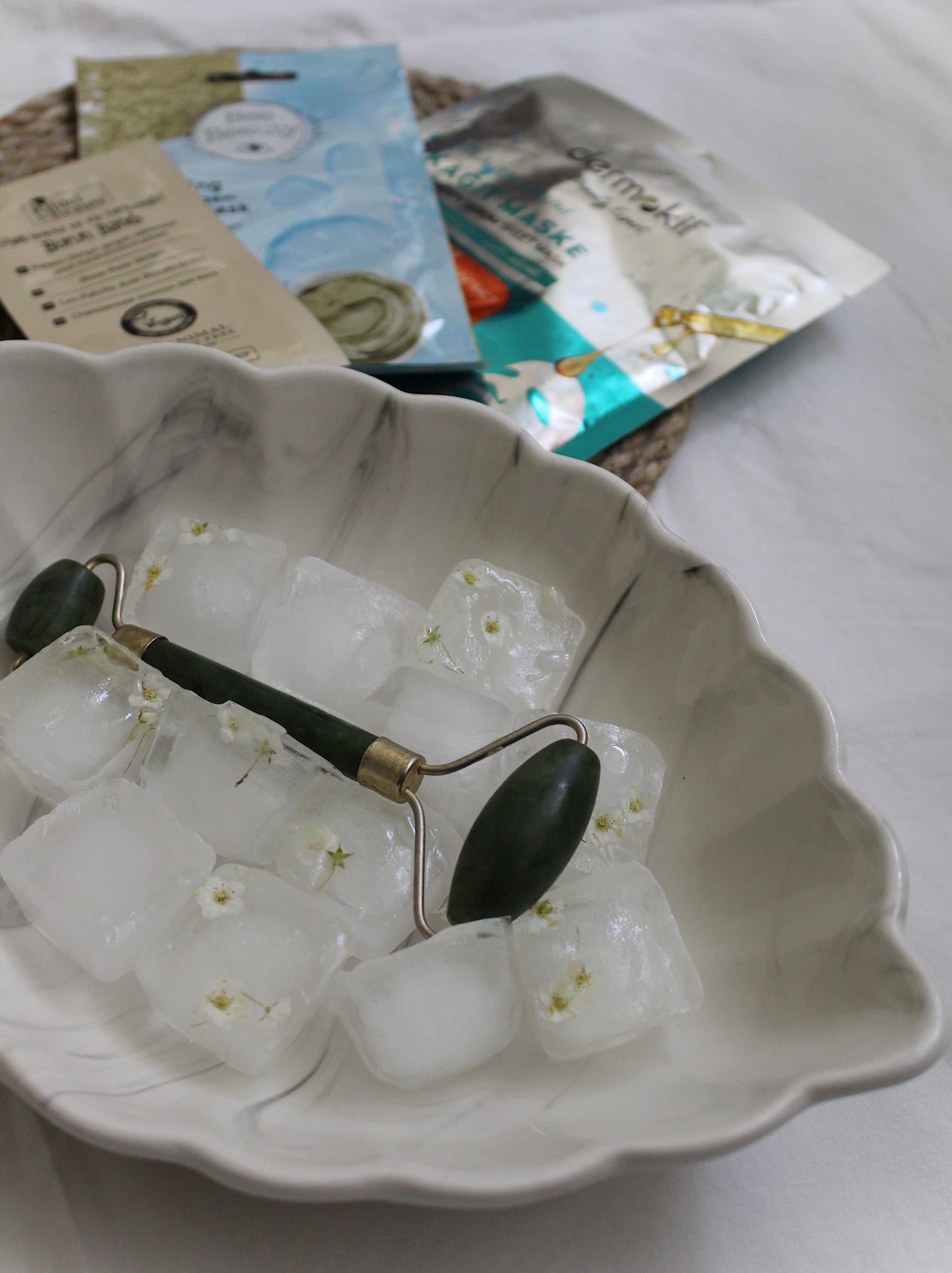 Jade Roller and Chilled Ice Cubes Skincare Jade roller with black handle resting on ice cubes infused with chamomile flowers in a white ceramic dish, with skincare product packaging in the background.