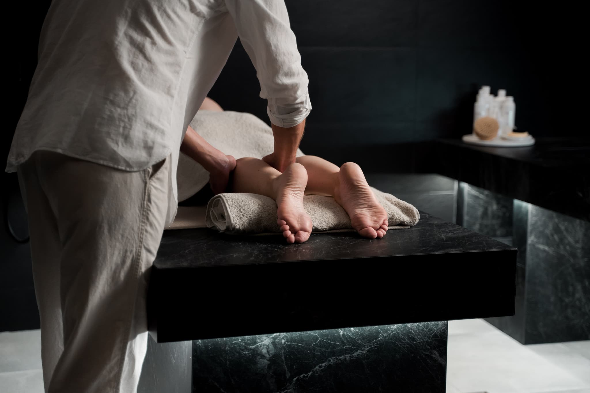 Foot Massage Spa Treatment Massage therapist performing a foot massage on a client lying on a black spa table in a dimly lit treatment room.
