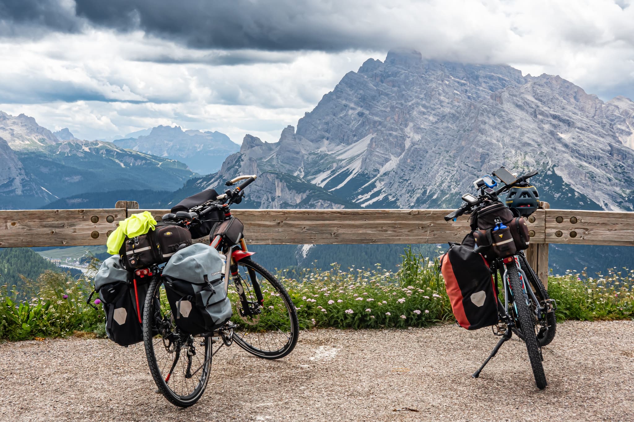 Tourenräder am alpinen Bergaussichtspunkt Zwei beladene Tourenräder parken an einem Holzgeländer mit Blick auf ein dramatisches alpines Tal mit gezackten Bergspitzen und bewaldeten Hängen.