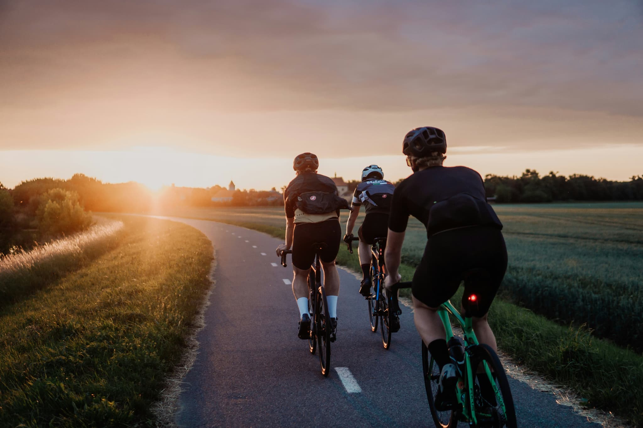 Cyclists on country road at sunset Three cyclists riding on a country road at sunset with green fields and a distant village.