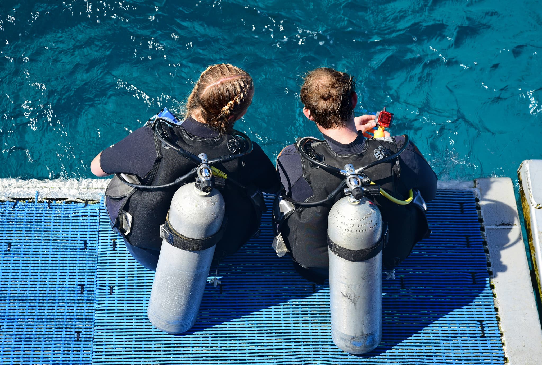 Divers Preparing on Boat Platform Two divers in black wetsuits sitting on a blue boat platform with air tanks, preparing to enter turquoise water.