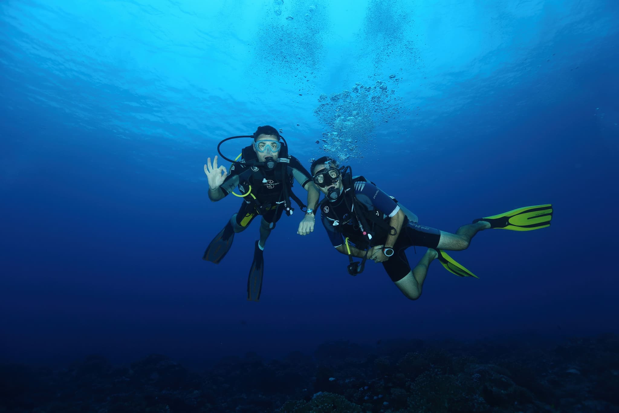 Scuba Divers in Blue Ocean Water Two scuba divers in wetsuits and gear swimming underwater in clear blue ocean water with bubbles rising above.