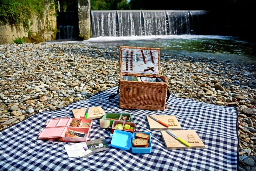 Riverside Picnic Spread by a Weir Picnic basket and bento boxes on a checkered blanket by a river weir.