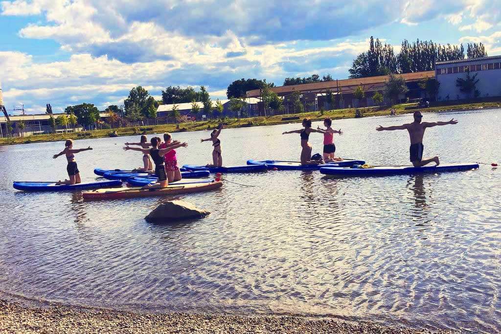 Paddleboard Yoga Class on Water Group of people practicing yoga poses on paddleboards in shallow water with a sandy beach and waterfront buildings in the background.