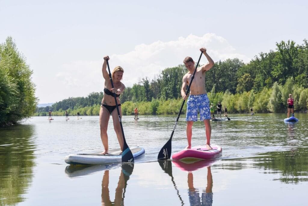 Paddleboarder on the river with mountain view Two paddleboarders on a calm river with paddles raised, surrounded by green trees and distant mountains.