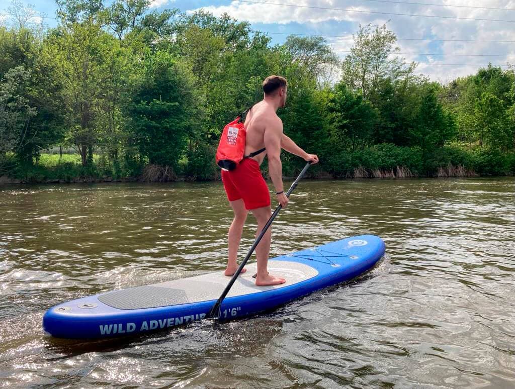 Paddleboarder on the river wearing a life jacket Person paddleboarding on a river, wearing a red life jacket and red shorts and holding a paddle.