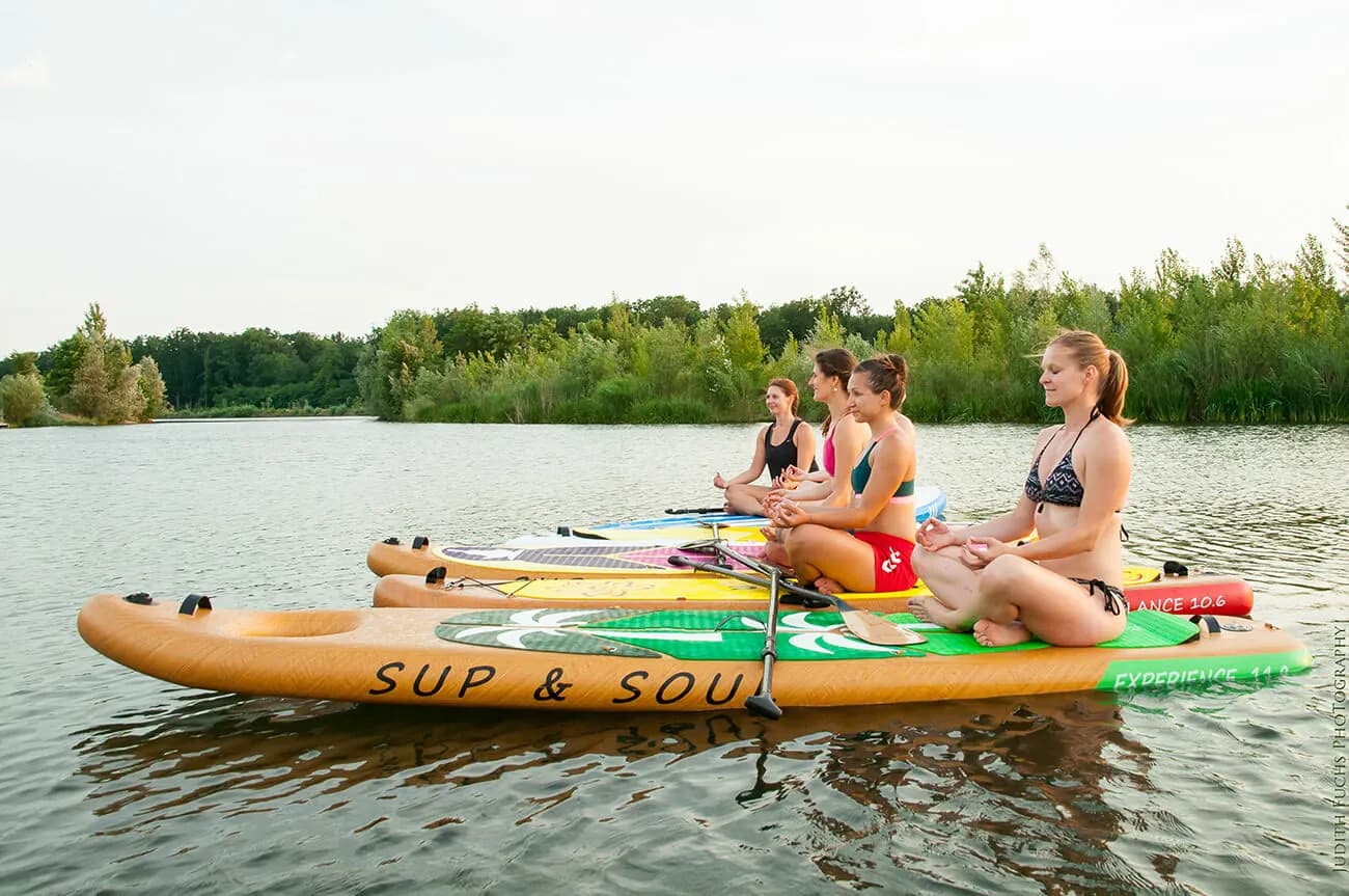 Paddleboarder on the Lake with Forest Four people sitting on paddleboards in calm lake water with a forested shoreline in the background.