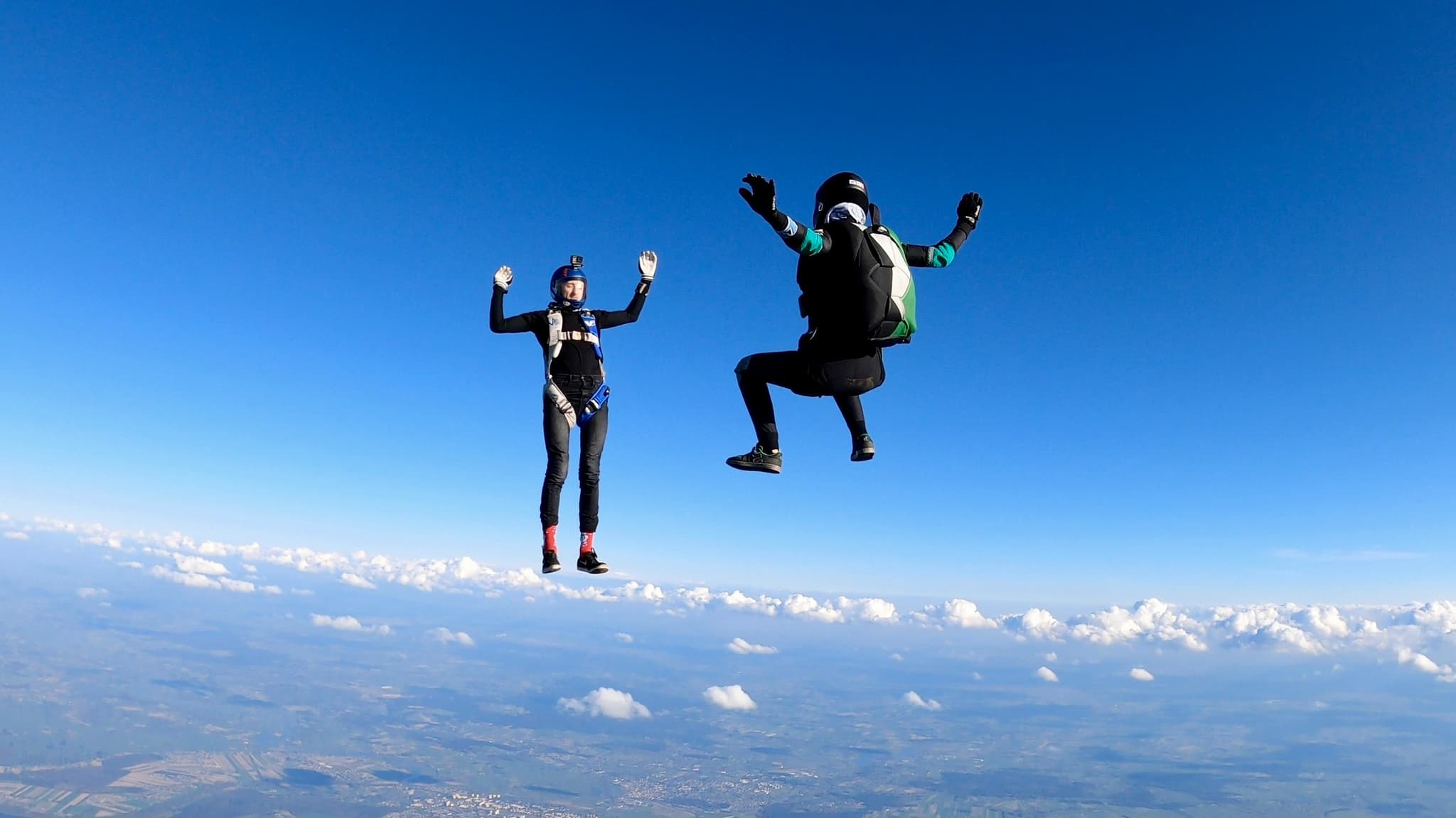 Skydivers celebrating in freefall Two skydivers in freefall against a blue sky with clouds and landscape below, both with arms raised in celebration