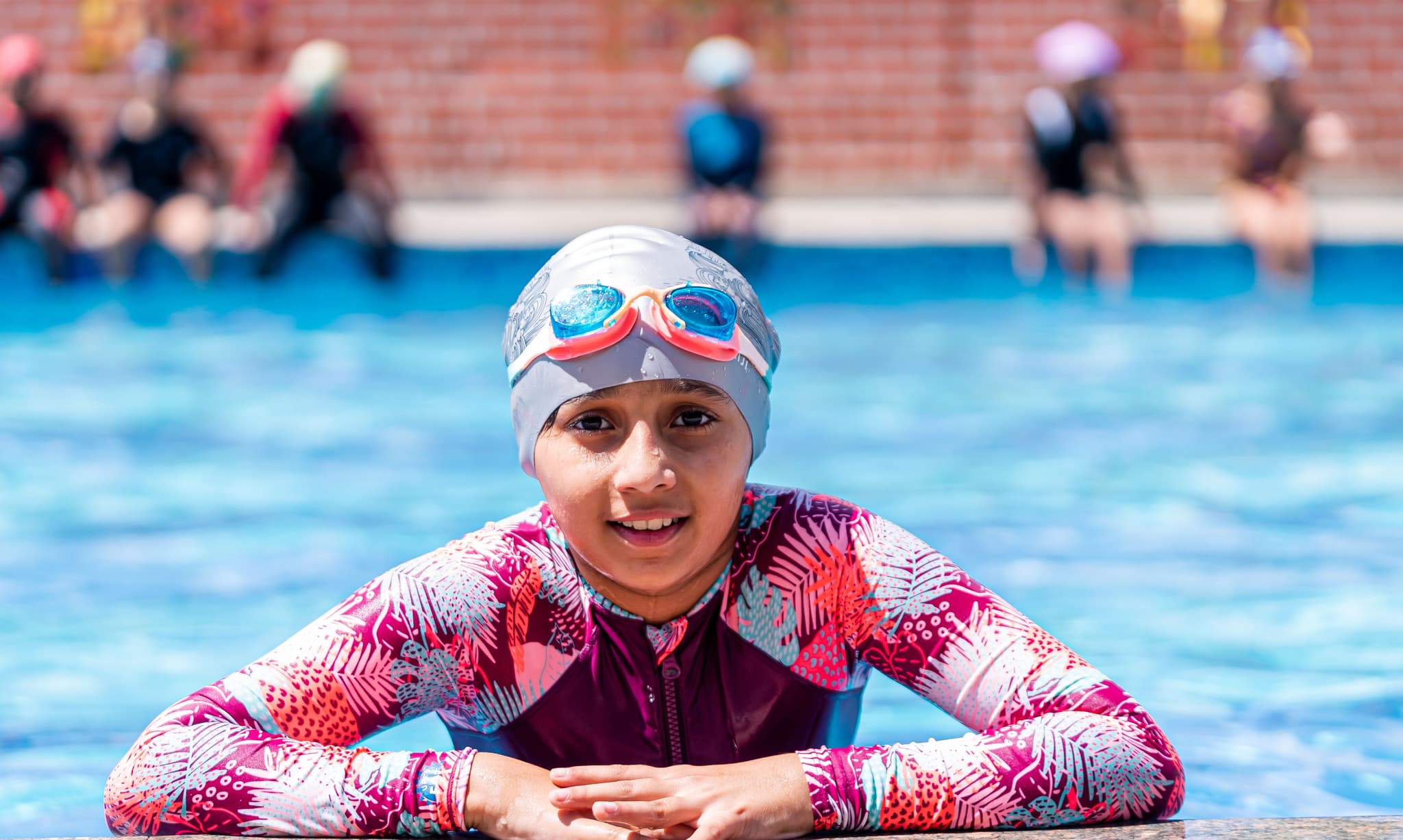 Child in swimming pool with swim goggles Child in a swimming pool wearing a white swim cap, red and blue swim goggles, and a colorful pink patterned swim shirt, smiling