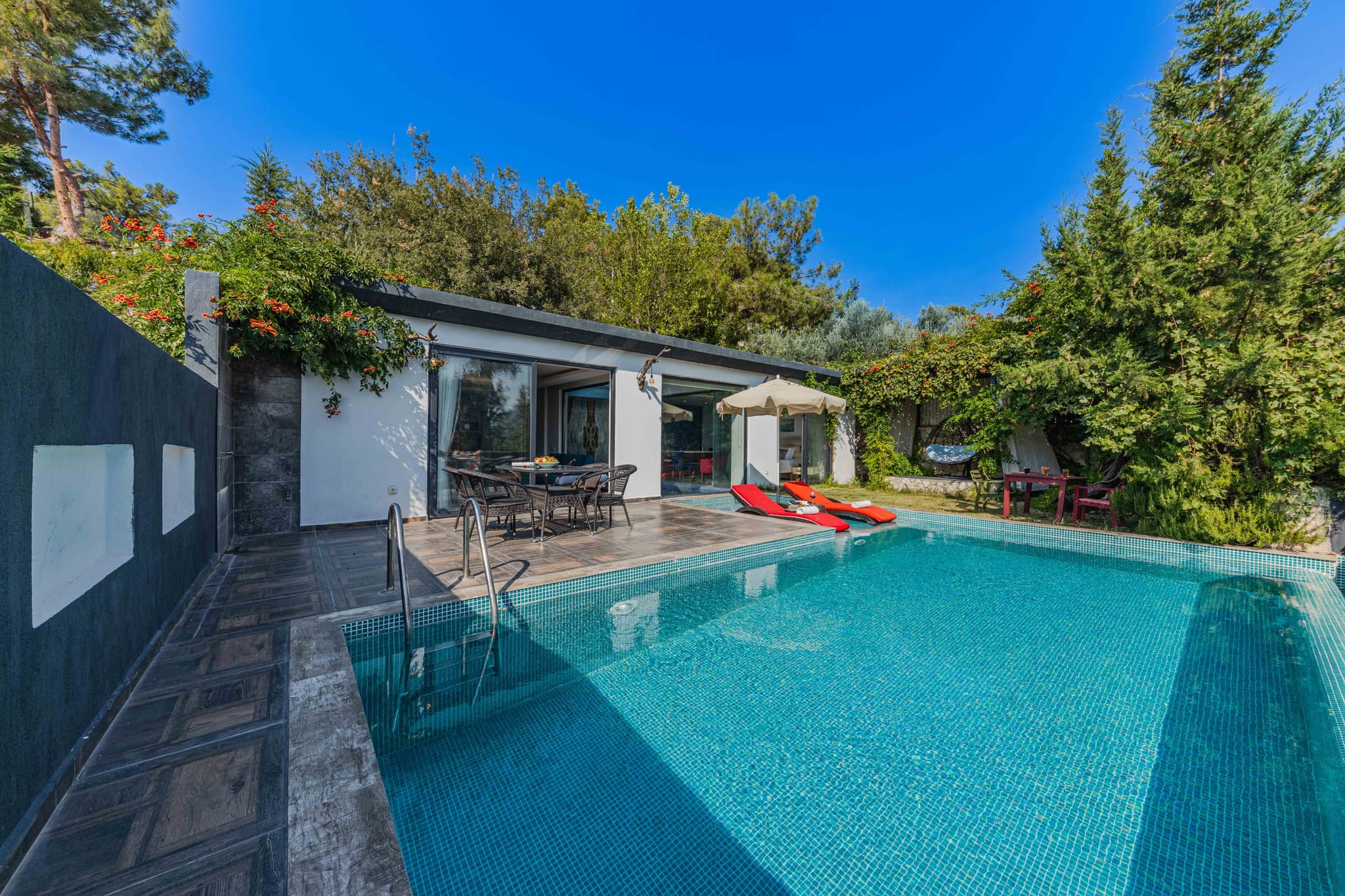 Modern Pool House With Terrace and Garden Modern pool house with turquoise swimming pool, red lounge chairs on deck, and surrounding evergreen trees under clear blue sky.