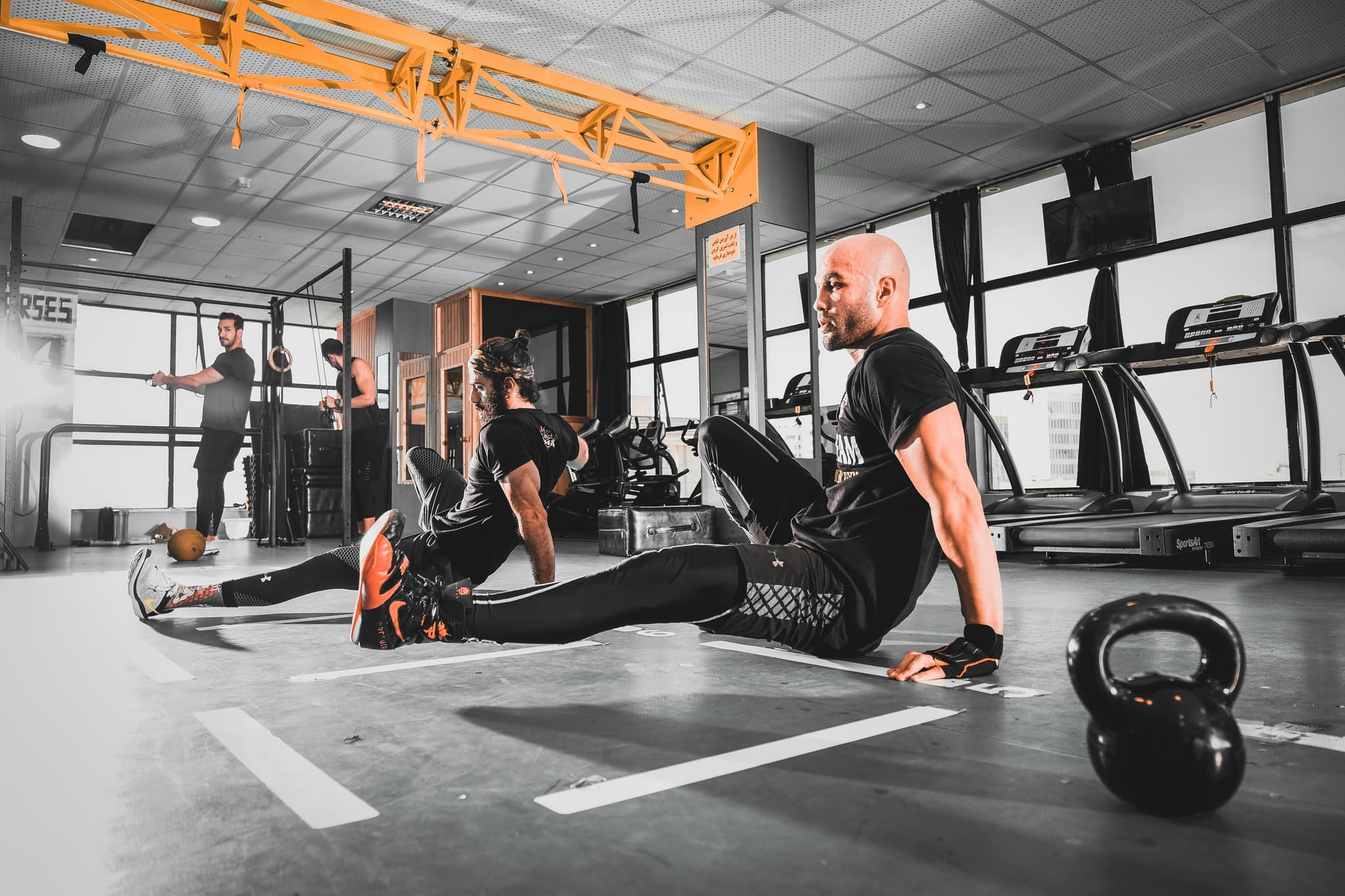 Gym Floor Stretch With Kettlebell Man stretching on gym floor near a kettlebell, others exercising in background.