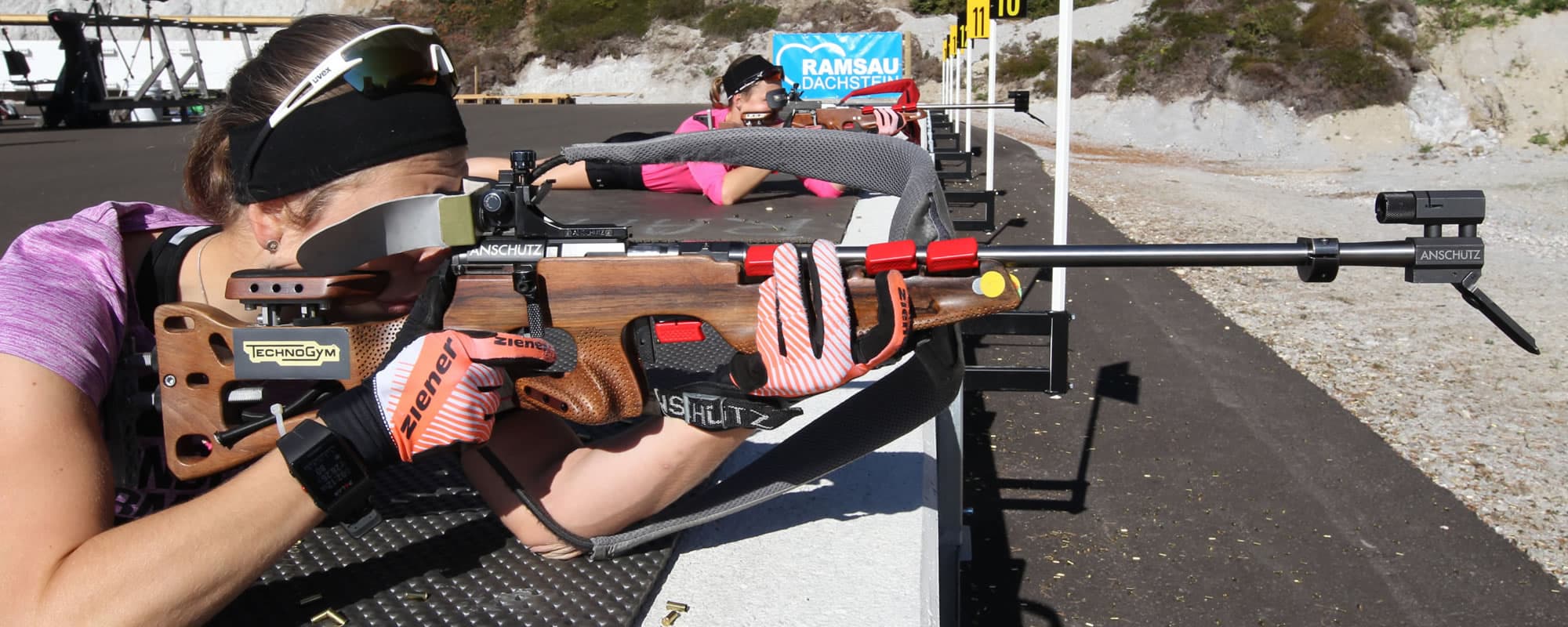 Precision Rifle Shooting Competition Shooter in pink and purple gear aiming a precision rifle at an outdoor shooting range with targets and hillside backdrop.