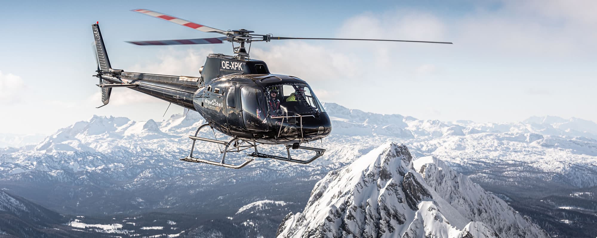 Helicopter Over Alpine Mountains Black helicopter in flight over snow-covered alpine peaks and forested valleys.