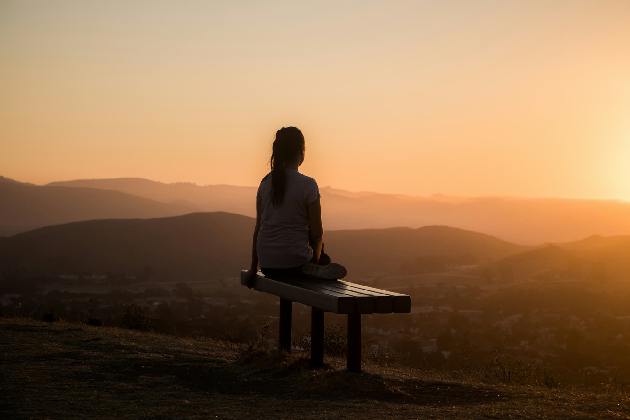 Sunset Mountain Valley Viewpoint Person sitting on a wooden bench overlooking rolling mountains at sunset with golden light across the valley.