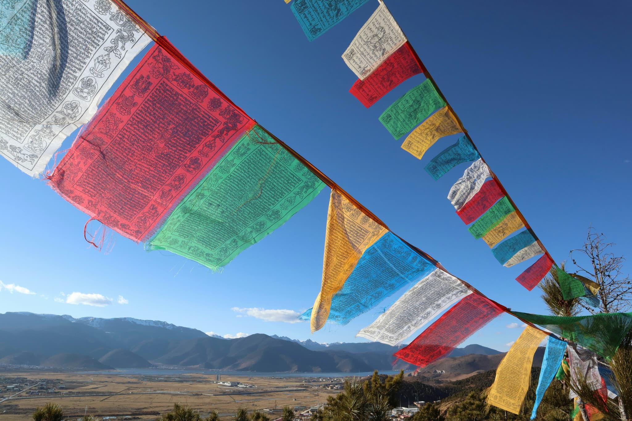 Prayer Flags Over Mountain Valley Colorful prayer flags strung across a mountain valley with snow-capped peaks and rural settlements in the distance.
