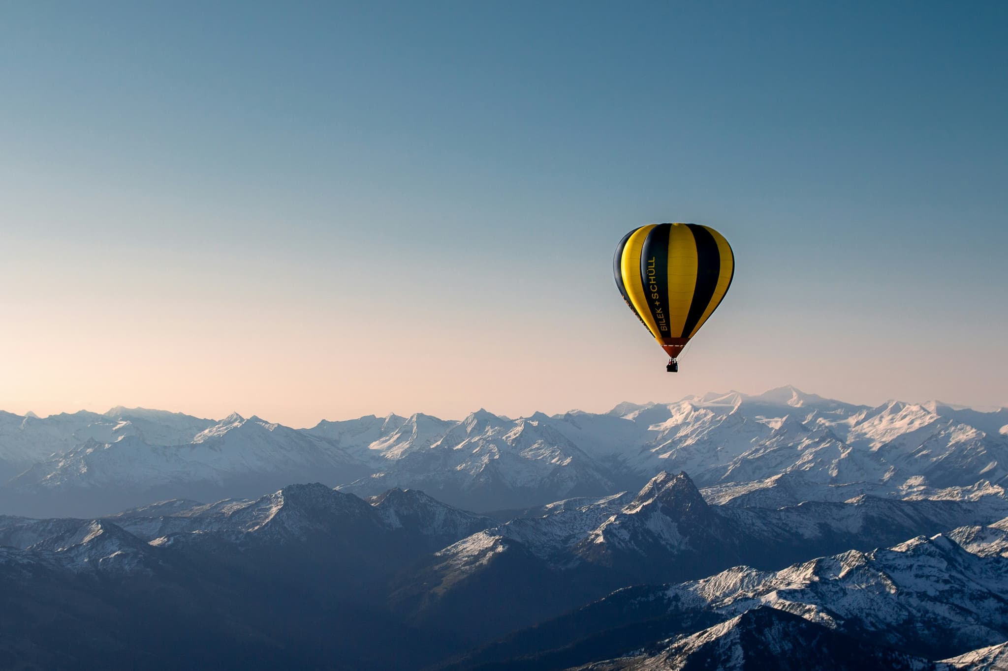 Hot Air Balloon Over Snow-Covered Mountains A hot air balloon with yellow and black stripes floats above a wide mountain range covered in snow under a clear blue sky.