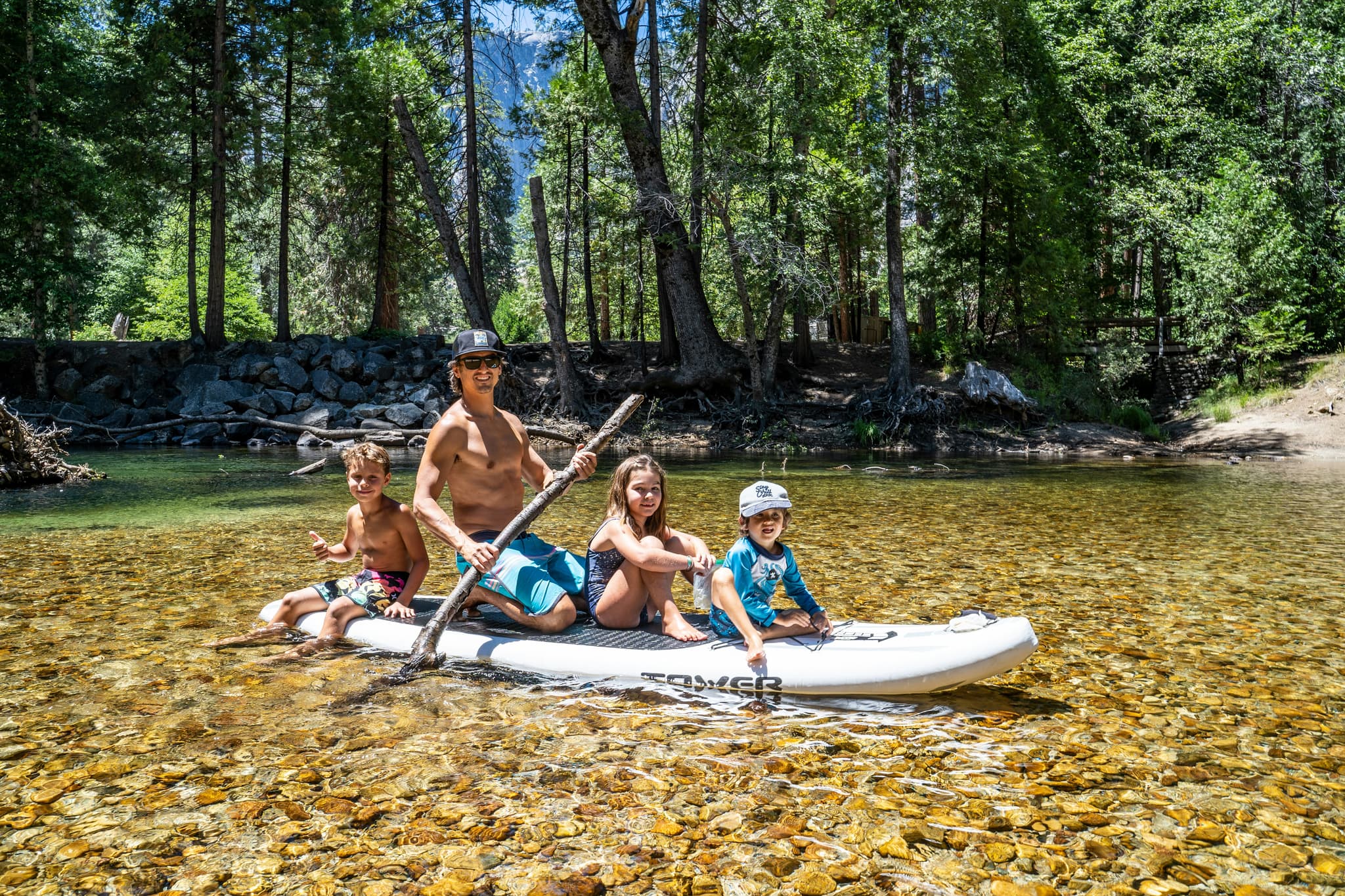 Family Paddleboarding on the Forest River An adult and three children on a paddleboard in a shallow river, surrounded by tall trees and mountains.
