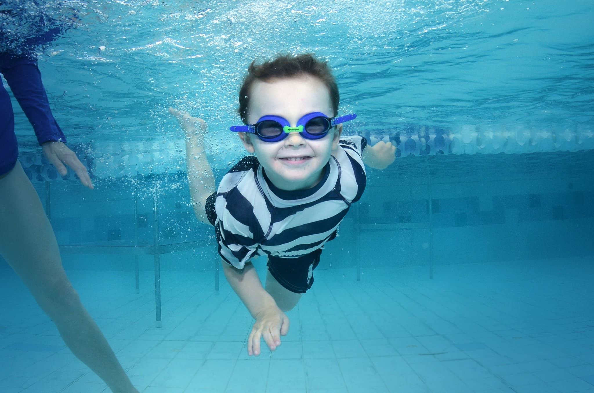 Child Swimming Underwater in Pool Child swimming underwater in a pool wearing blue goggles and a striped swim shirt.