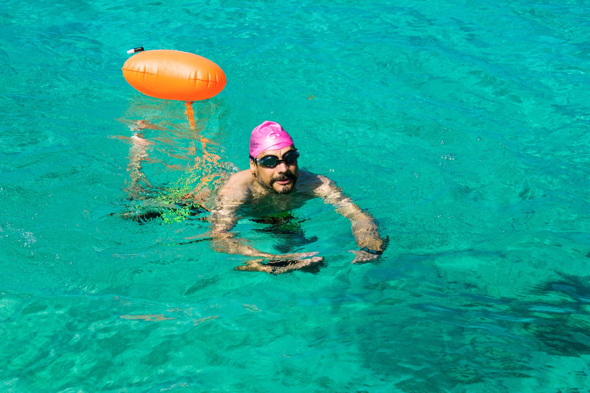 Schwimmer mit orangefarbenem Rettungsring Schwimmer in rosa Badekappe und Schwimmbrille beim Freistilschwimmen in klarem türkisblauem Wasser mit orangefarbenem Rettungsring in der Nähe.