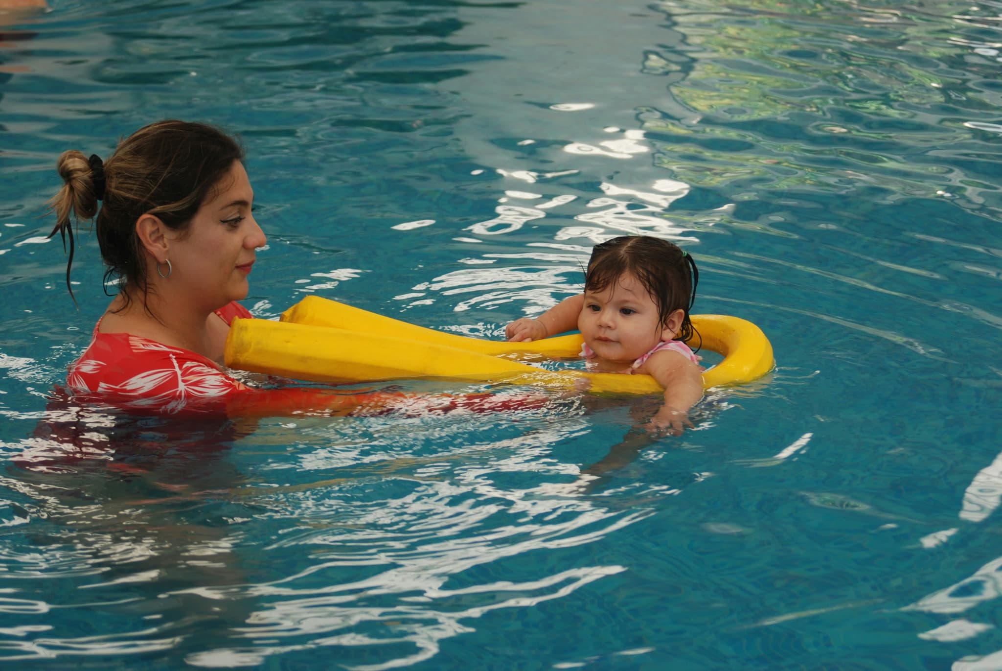 Schwimmunterricht für Kleinkinder mit Auftriebshilfe Eine erwachsene Person beaufsichtigt ein Kleinkind im Schwimmbecken mit einem gelben Auftriebsgerät.