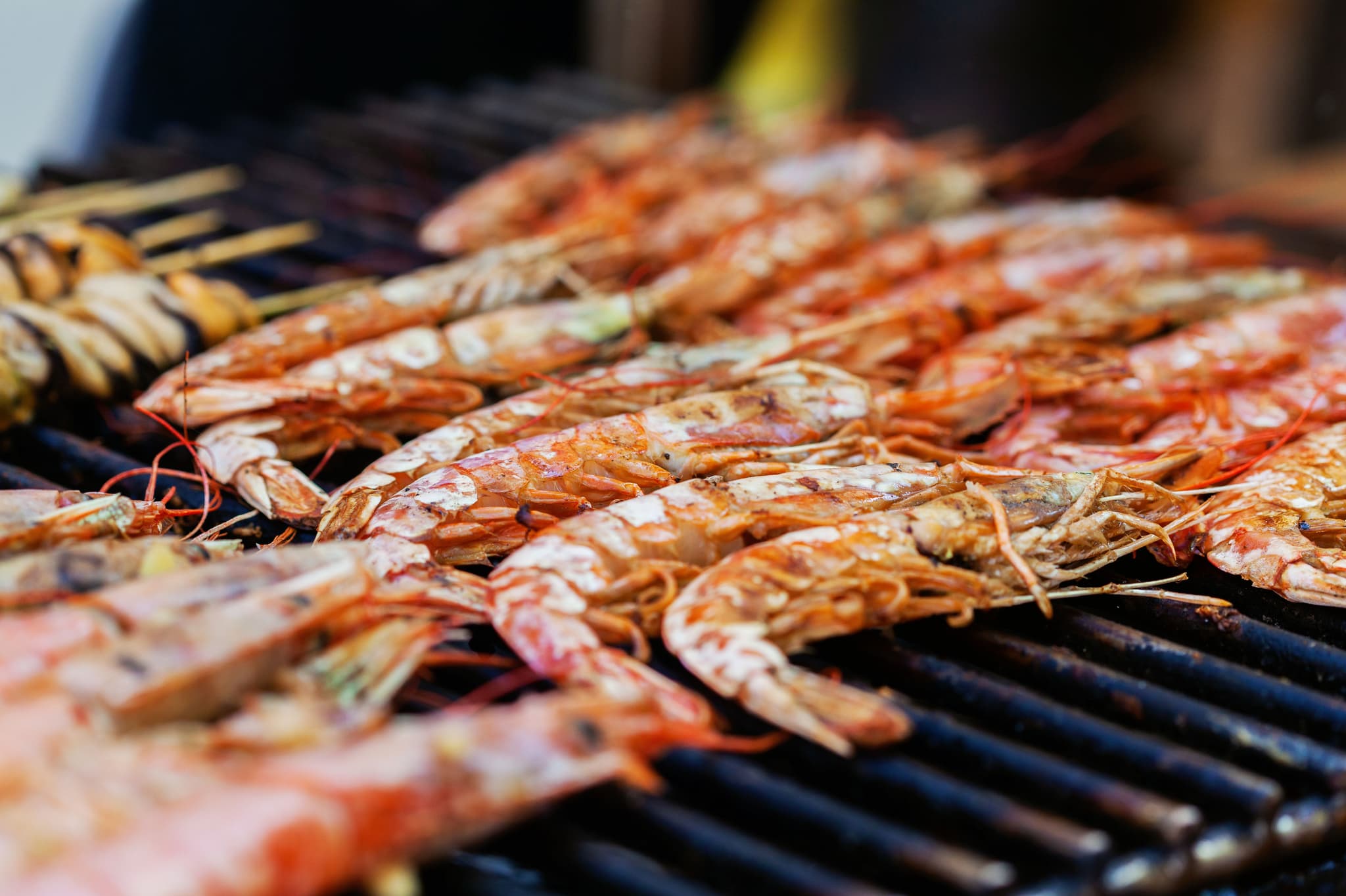Grilled Shrimp on Barbecue Grill Grilled shrimp on a barbecue grill with char marks and vegetables in the background.