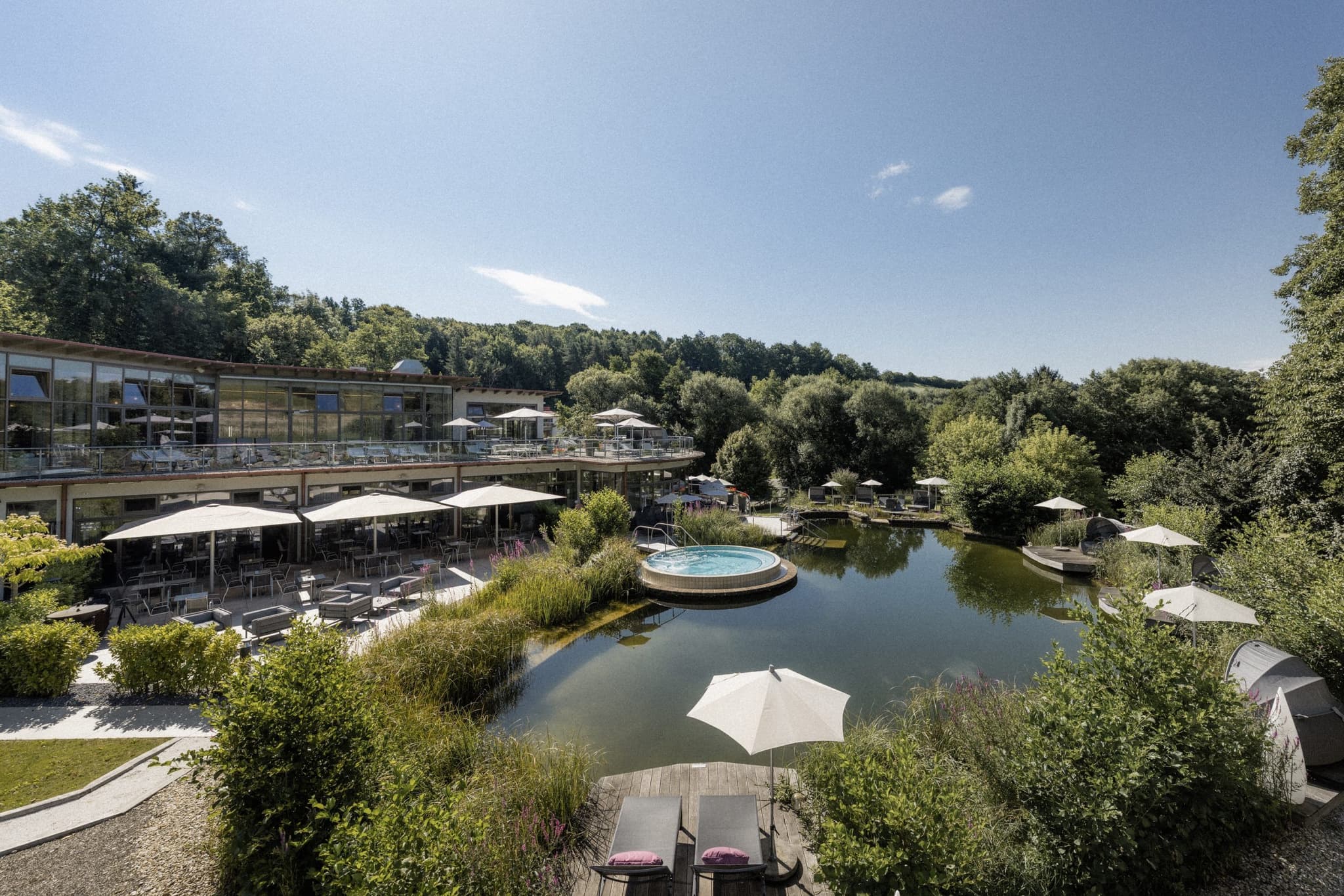 Spa Resort Pool and Terrace Woodland View Outdoor spa resort with circular hot tub, lounge seating, and dining terrace overlooking a pond and forested landscape.