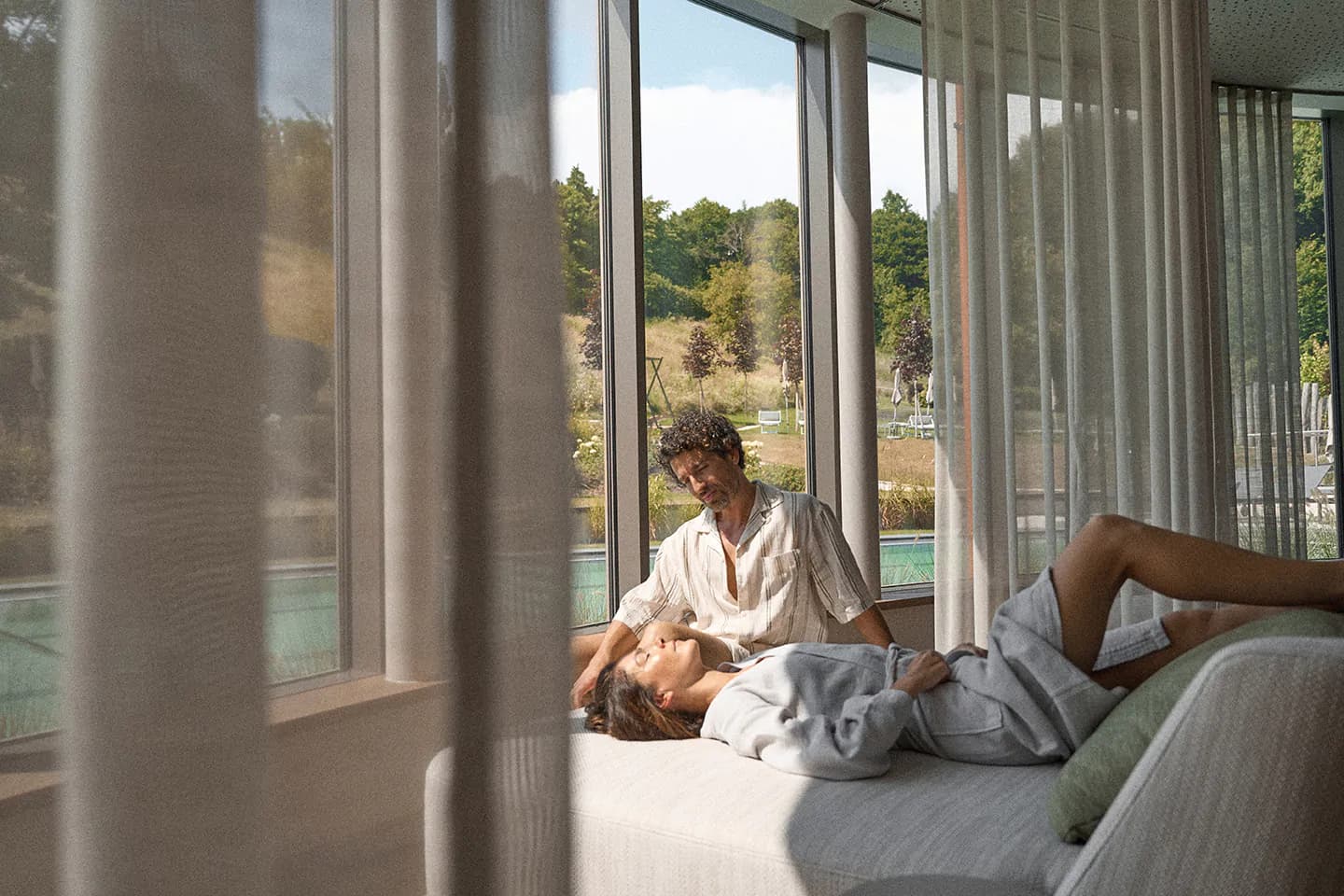 Couple Relaxing by Windows With Countryside View Two people relaxing on a bed by large windows overlooking a countryside landscape with trees and open fields.