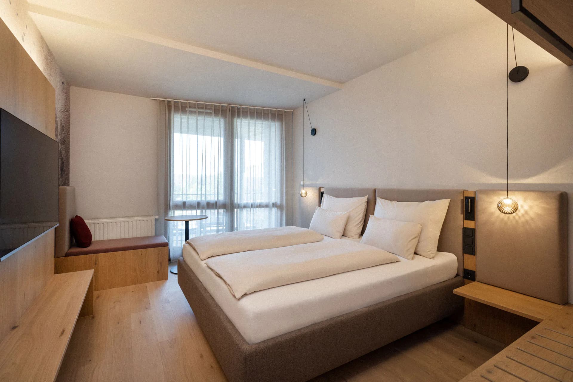 Modern Hotel Bedroom With Balcony Modern hotel bedroom with a gray upholstered bed, wooden furniture, sheer curtains, and a balcony with city views.