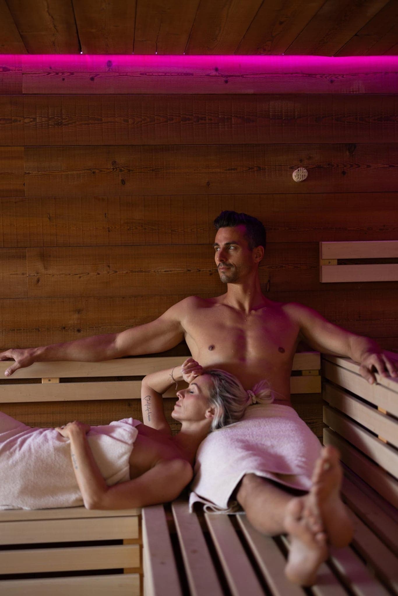 Couple in Sauna with Neon Lighting Two people relax together in a wooden sauna with pink neon accent lighting.
