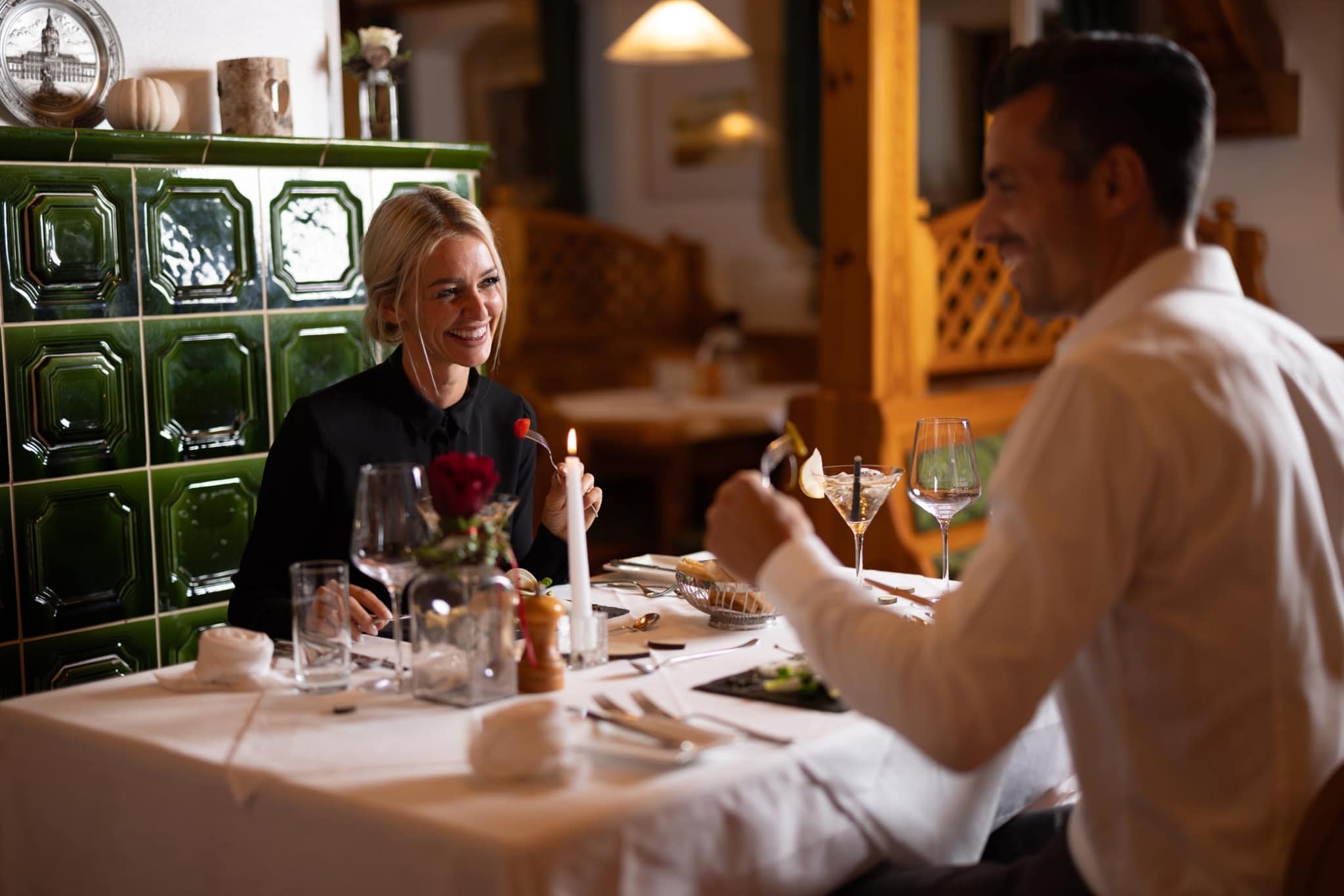 Elegant Restaurant Dining Experience Two people dining at an elegant restaurant table with wine glasses, a red flower centerpiece, and a green tiled wall backdrop.