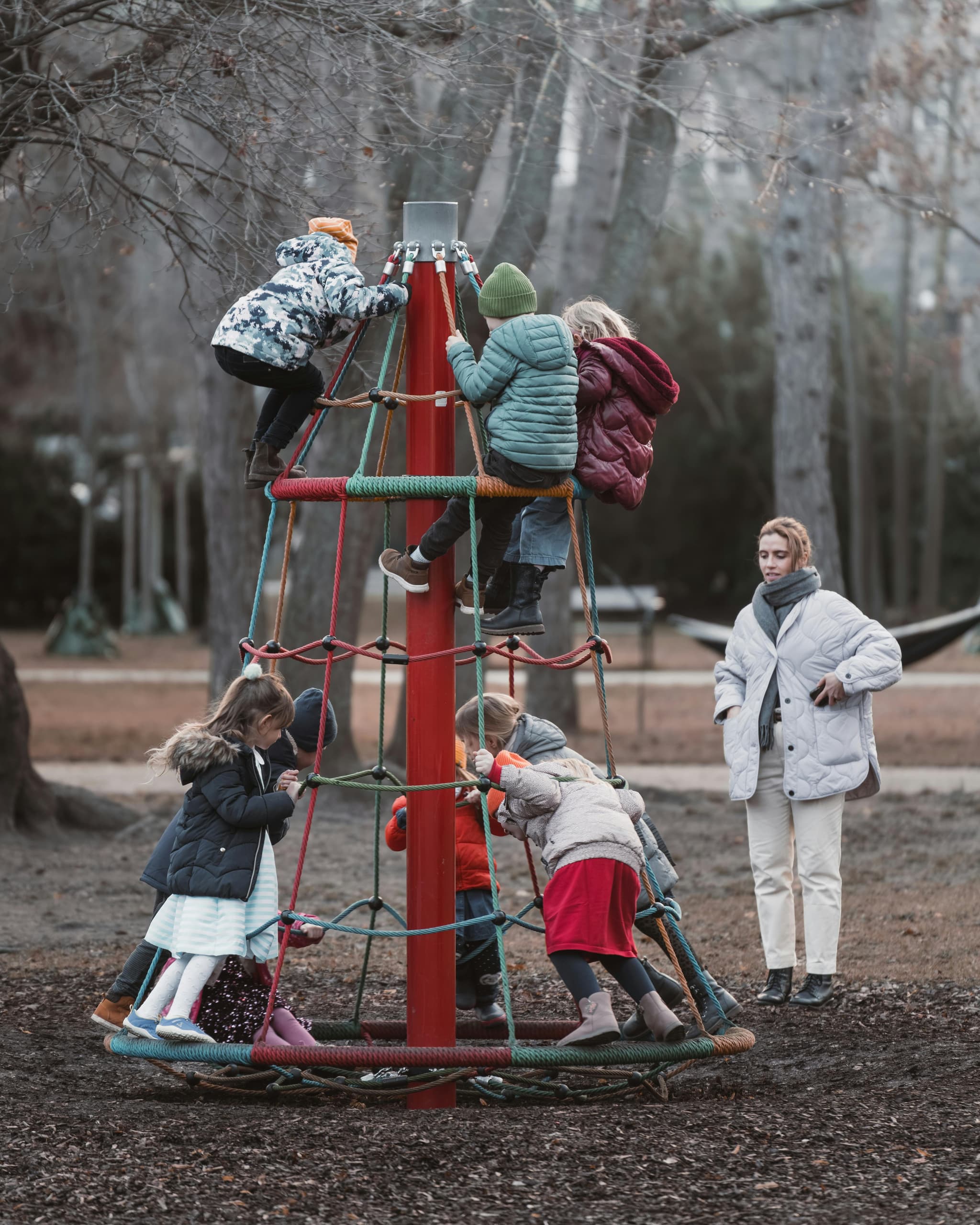 Children Climbing Rope Playground Structure Children climbing a rope pyramid structure in a park while an adult watches nearby.