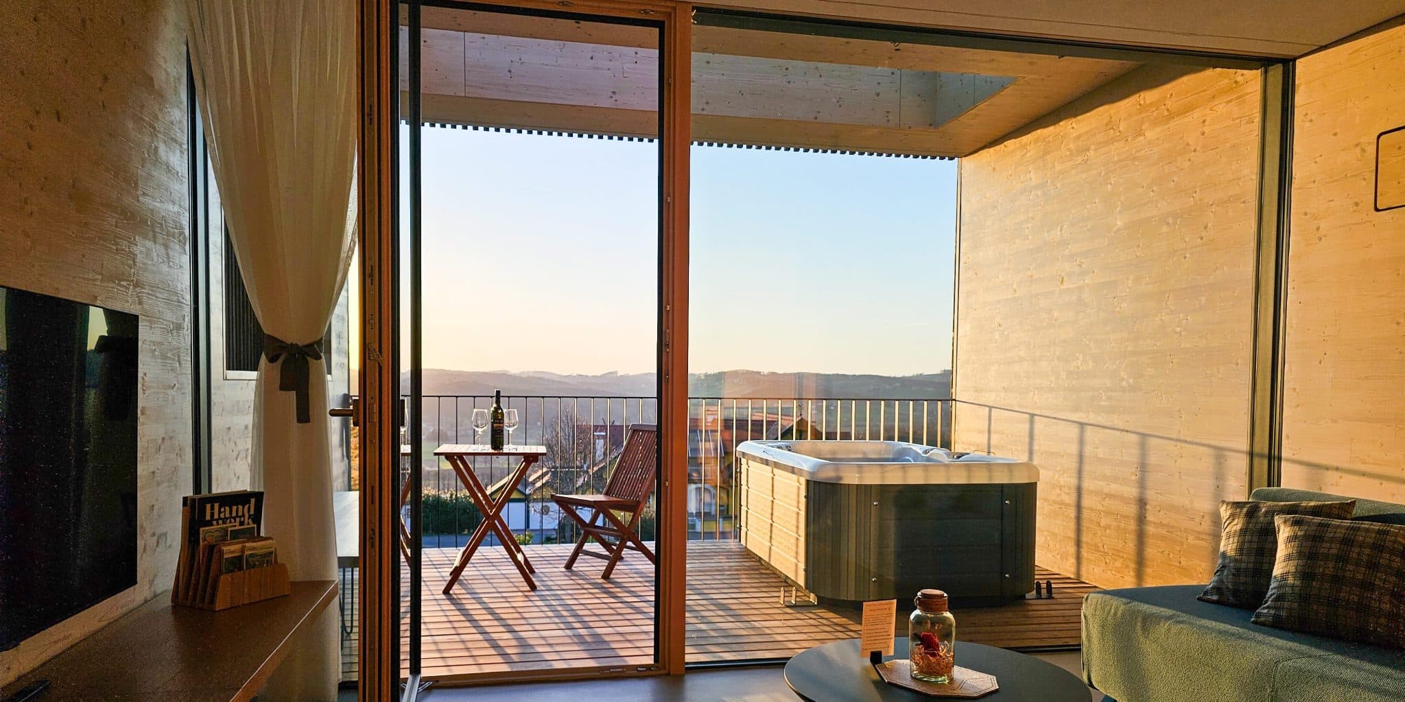 Hotelsuite mit Balkon, Whirlpool und Bergblick Moderne Hotelsuite mit offenen Glastüren zu einer Holzterrasse, Whirlpool und Bergblick bei Sonnenuntergang.