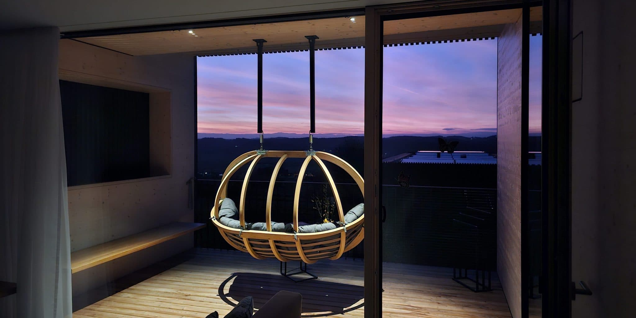 Hängende Pod-Sessel auf moderner Terrasse bei Sonnenuntergang Holzerner hängender Pod-Sessel mit Kissen, aufgehängt von einer modernen Terrasse mit Blick auf Berge bei Sonnenuntergang.