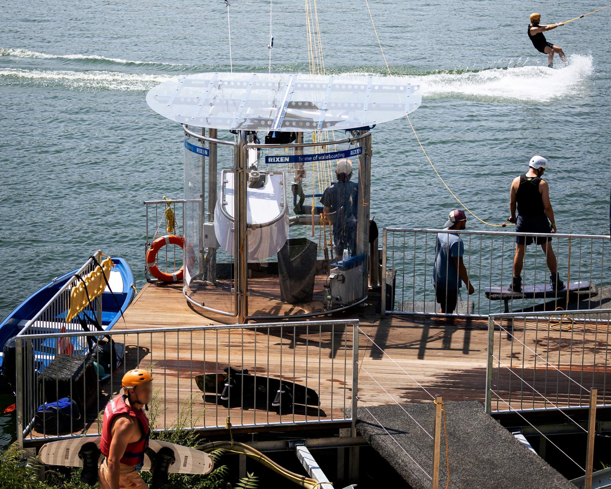 Wakeboarding Boat and Water Sports Dock Wakeboarding boat docked at a wooden pier with wakeboarders and equipment nearby, ocean waves in the background.