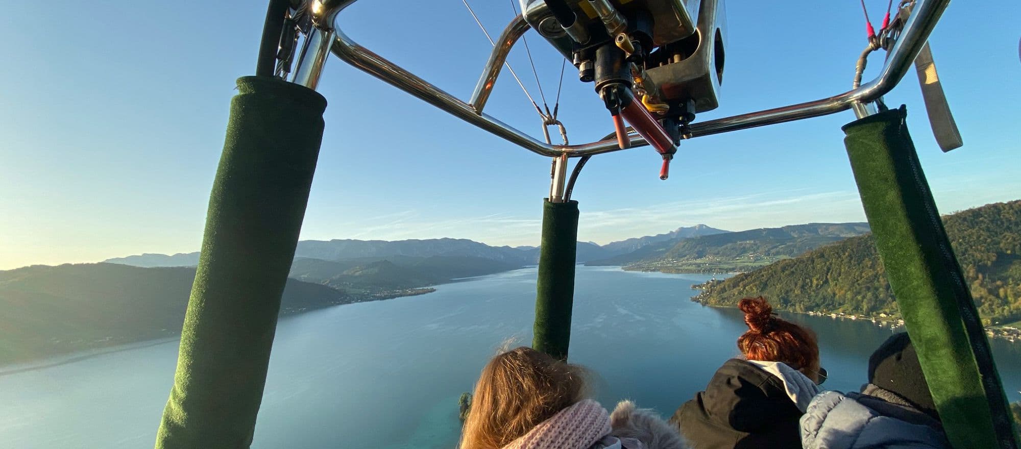 Paragliding Over Alpine Lake and Mountains Aerial view from a paraglider showing two people suspended above a pristine alpine lake surrounded by forested mountains and scattered settlements.