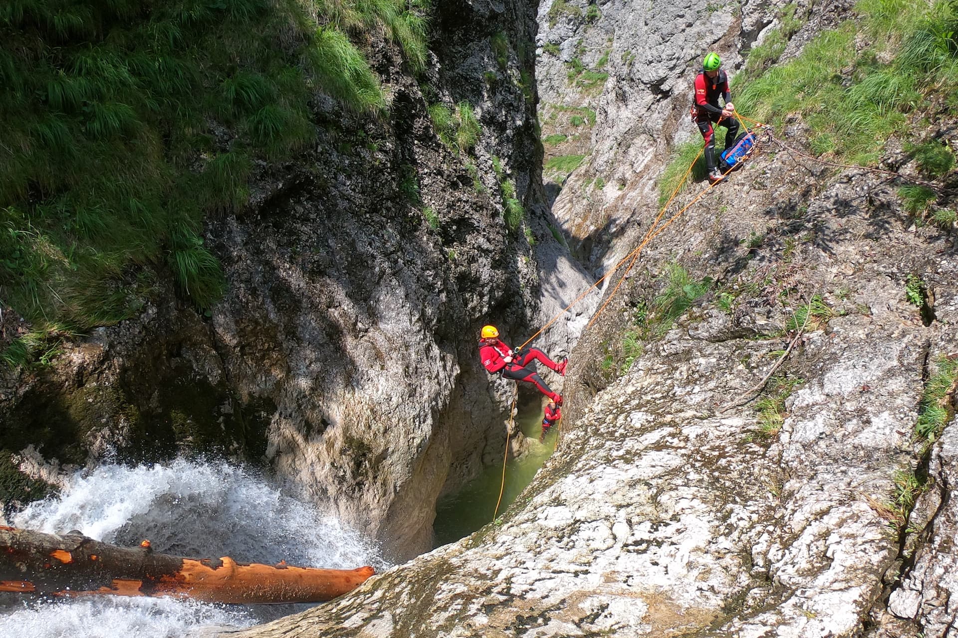 Canyoneering Rappel in Rocky Gorge Person rappelling down a rocky canyon wall above a flowing stream, with another participant visible on the slope above.