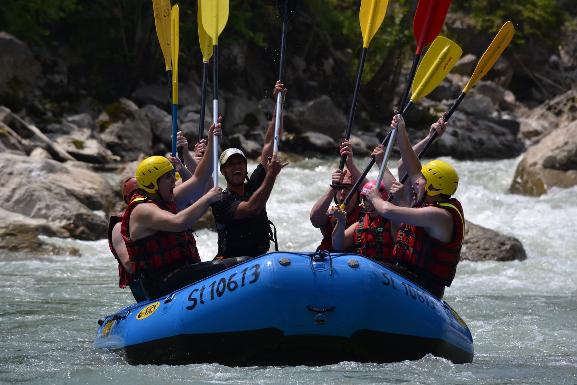 Whitewater Rafting Group on River Group of people whitewater rafting on a river, paddling together in a blue raft with yellow and red paddles.