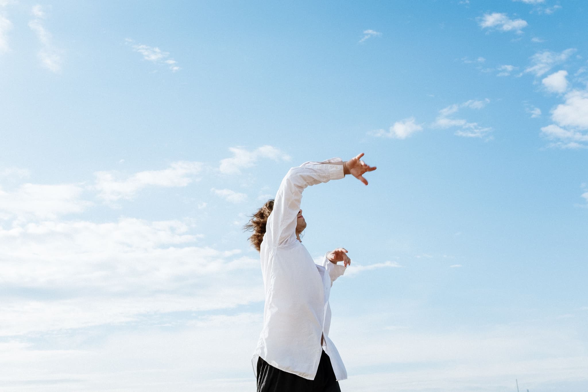 Person Stretching Outdoors Under Blue Sky Person in white shirt stretching arms upward against a bright blue sky with white clouds.