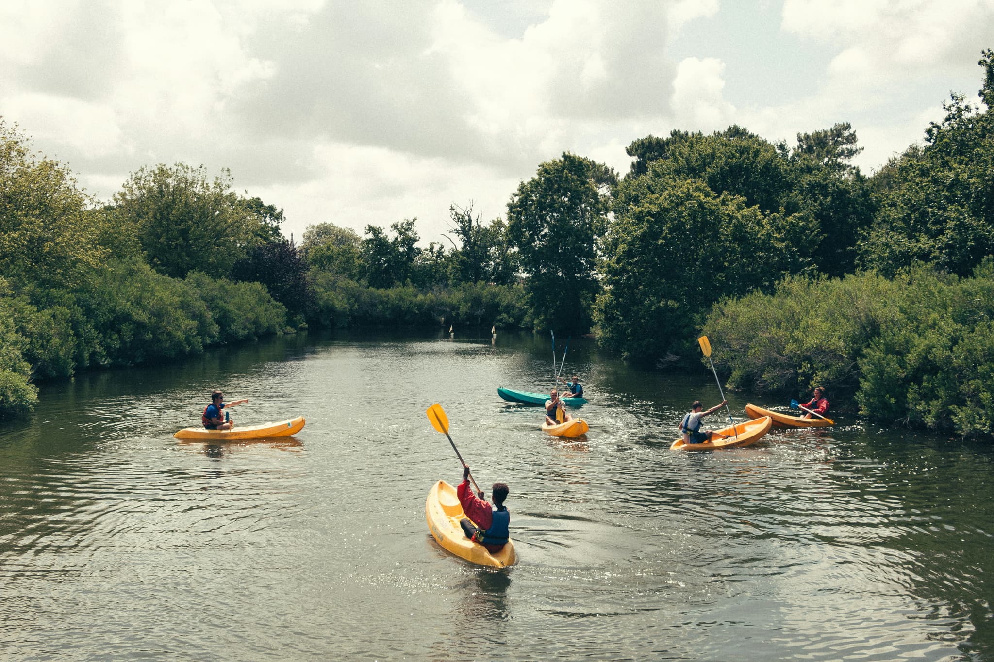 Group kayaking adventure on the river Multiple people paddling in yellow and turquoise kayaks on a calm river, surrounded by dense green forest.