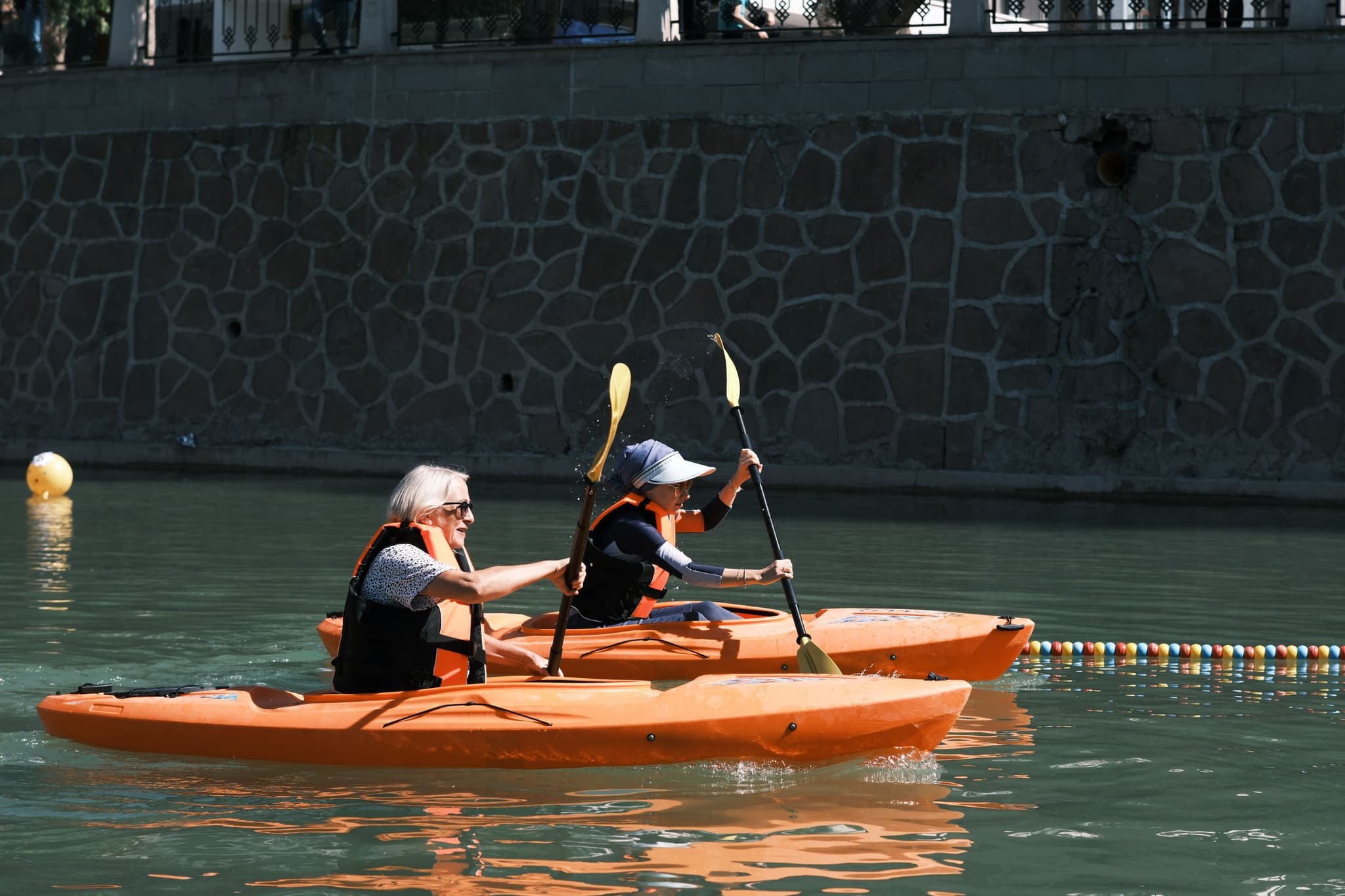 Kayaking on Urban Waterway Two people paddling orange kayaks on calm water with a stone wall and building in the background.