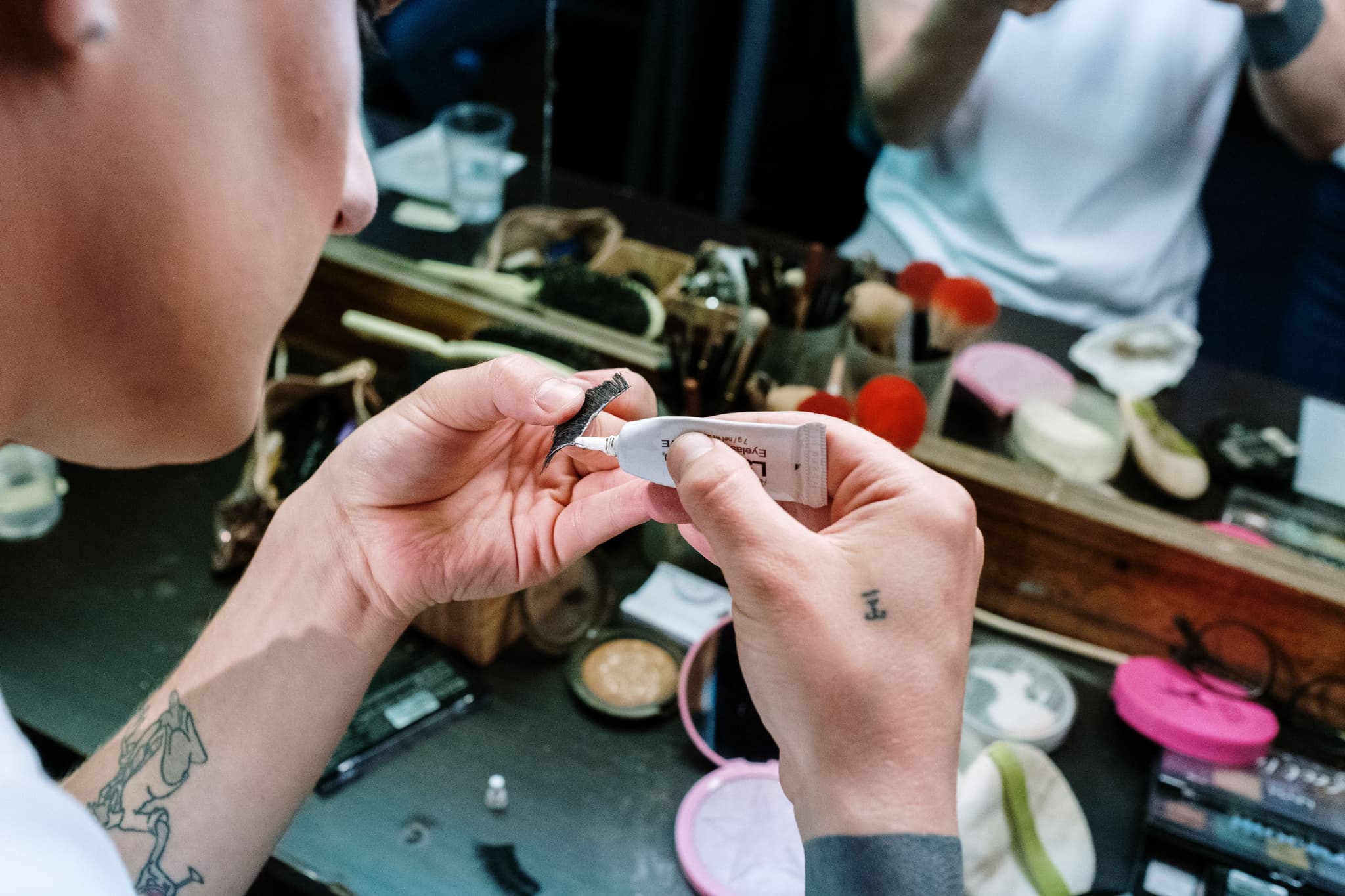 Makeup Product Examination at Vanity Person holding a makeup product and examining it at a vanity table with mirrors, surrounded by cosmetic products.